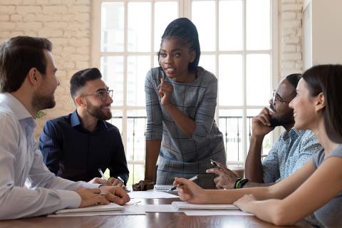 People at conference table smiling at standing person in center.